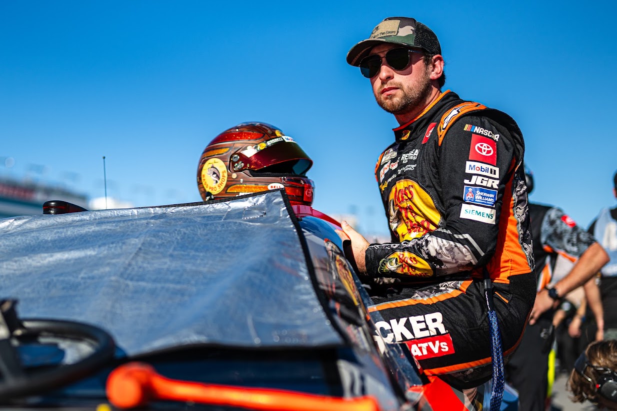 Chase Briscoe in his Bass Pro Shops firesuit, camo cap and sunglasses, beside the covered No. 19 Toyota on the grid